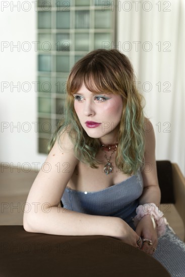 A woman with colorful, pastel hair leans thoughtfully on a brown table Dressed in a light blue