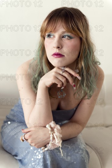 A woman with green-tinted hair and expressive makeup is seated on a white couch She wears a blue