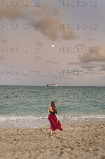 Back view of an unrecognizable redhead woman strolling along Miami Beach. She wears a flowing red