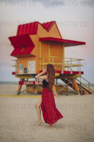 Back view of unrecognizable redhead woman walking near a vividly colored lifeguard station on Miami