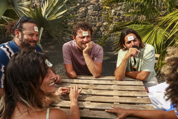 A multiethnic group of friends plays a guessing game at a wooden table outdoors They laugh while