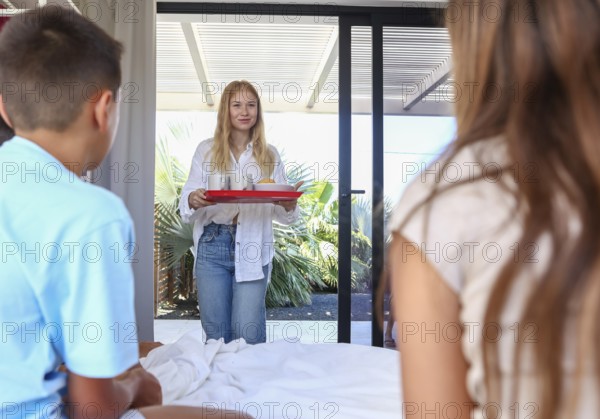 An older sister carrying a tray with drinks smiles as she serves breakfast to her younger siblings,