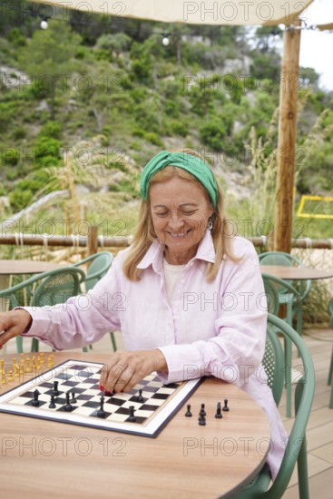 A senior woman plays chess outdoors on a sunny day, seated at a round table surrounded by greenery