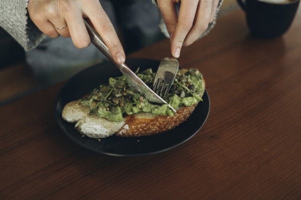 Close-up of hands using a fork and knife to slice avocado toast on a dark plate. The open-faced