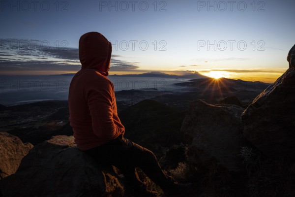 Back view of an unrecognizable man sitting on a mountain peak, watching a stunning sunrise over a