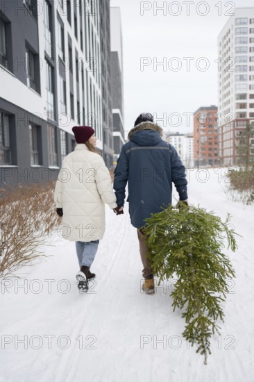 A couple walks hand in hand through a snow-covered urban area, carrying a freshly cut Christmas fir
