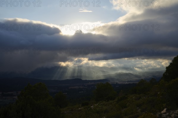 A breathtaking view of sunlight piercing through thick clouds over rolling hills and valleys,
