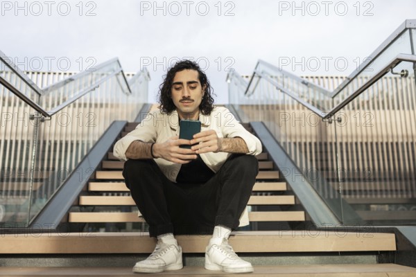 Young man sitting on outdoor stairs, casually browsing his smartphone The setting is urban and