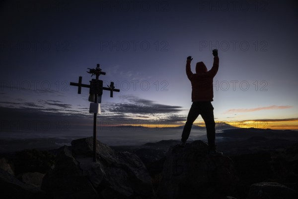 Unrecognizable man stands with arms raised near a silhouetted cross at sunrise, atop a rugged