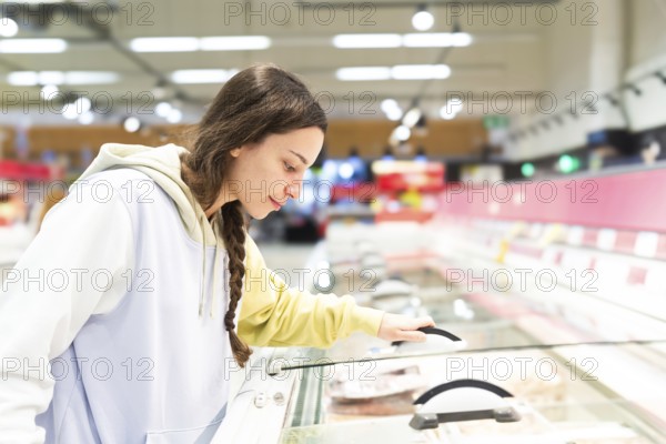 Side view of a woman is observed closely examining the products in a supermarket freezer section,