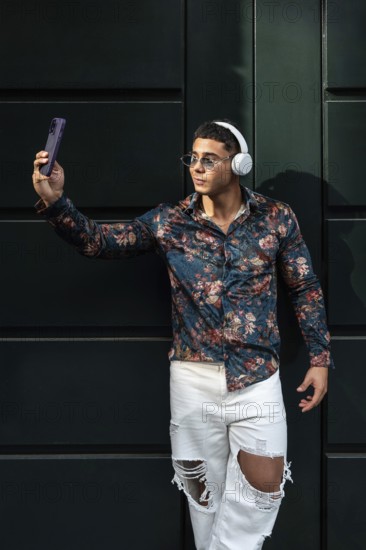 Stylish man wearing floral shirt and headphones takes a selfie. He wears sunglasses and ripped
