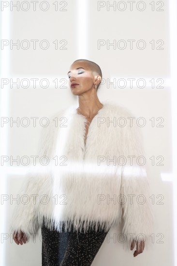 A middle-aged woman with a shaved head poses confidently, wearing a fashionable white faux fur coat