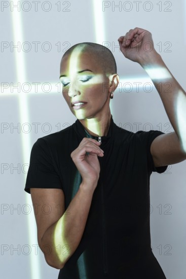A middle-aged woman with a shaved head poses gracefully under artistic light patterns, radiating