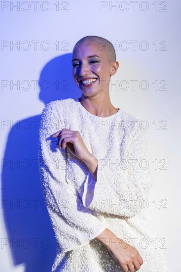 Middle-aged woman with a shaved head smiling confidently in a textured white outfit. Blue lighting