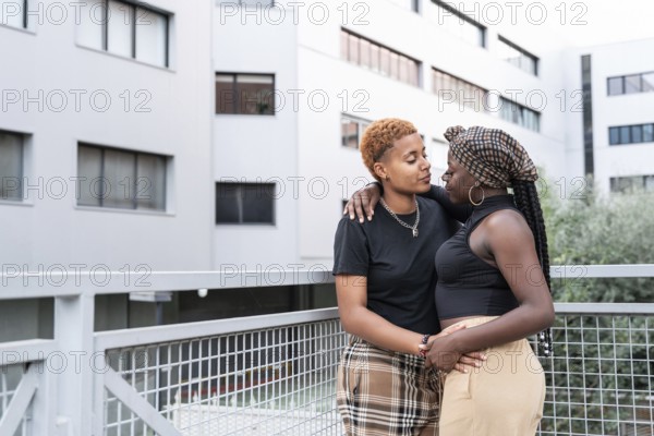 An affectionate LGBT multiethnic couple leans against a balcony railing in an urban setting,