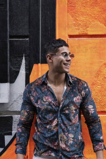 A stylish latin man in a floral shirt and sunglasses stands smiling against a vibrant orange and
