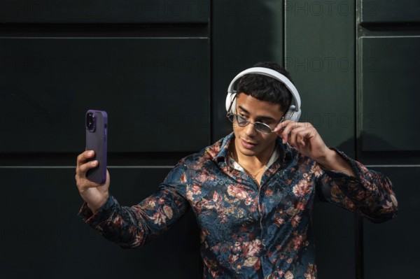 A trendy latin man in floral shirt and sunglasses takes a selfie outdoors, showcasing modern style