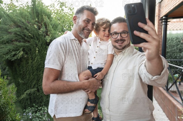 A joyful gay couple takes a smiling selfie with their son in a lush garden setting. The image