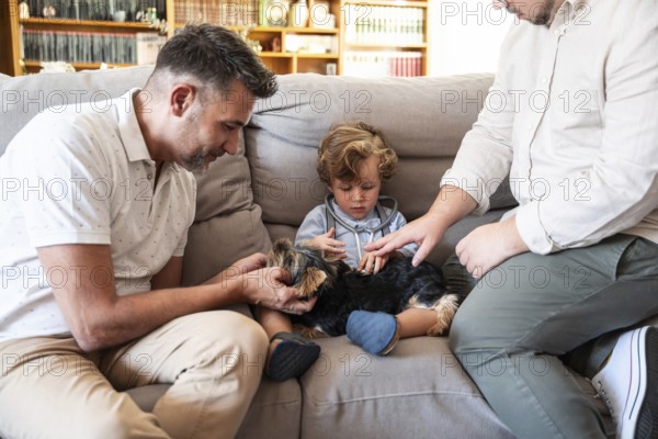 A loving gay couple interacts with their young son and small dog on a cozy couch in their living