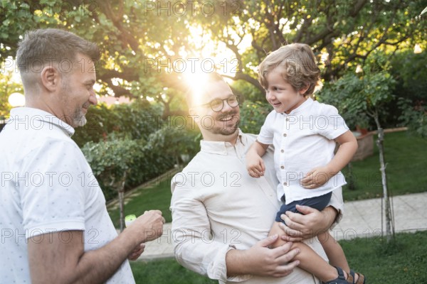 A joyful gay couple shares a tender moment with their son in a sunlit garden, surrounded by lush