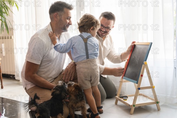 A joyful moment with a gay couple and their son drawing on a chalkboard, surrounded by playful dogs