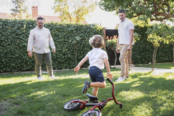 A loving gay couple shares a joyful moment with their young son playing with bicycle in the garden.
