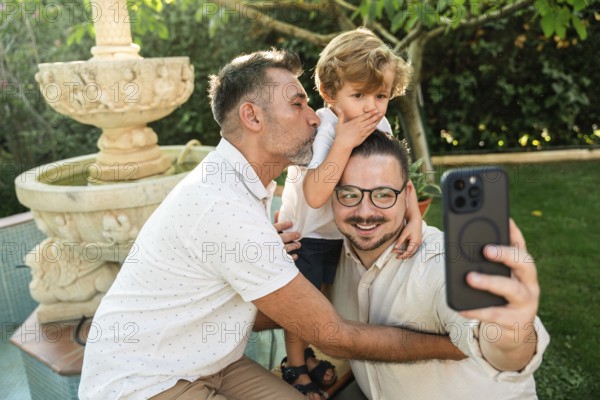A joyful gay couple takes a selfie with their young son in a lush garden. One dad kisses the son
