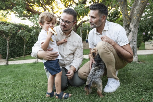 A joyful family moment in a lush garden with a gay couple watching their son eating a banana, and a