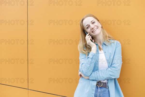 A cheerful woman stands against a bright orange wall in Warsaw, chatting on her phone She's dressed