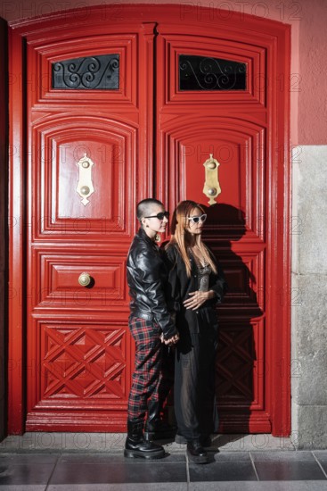 A fashionable lesbian couple stands lovingly against a bright red door, embracing each other's