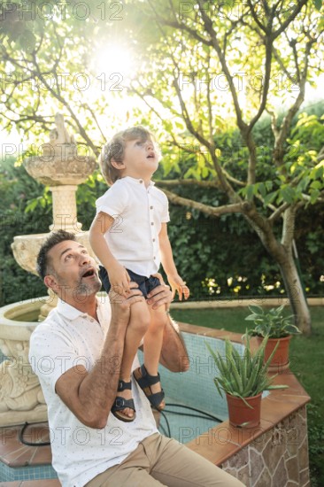 A father lifts his son as they gaze at something fascinating. Sunlight filters through the leaves