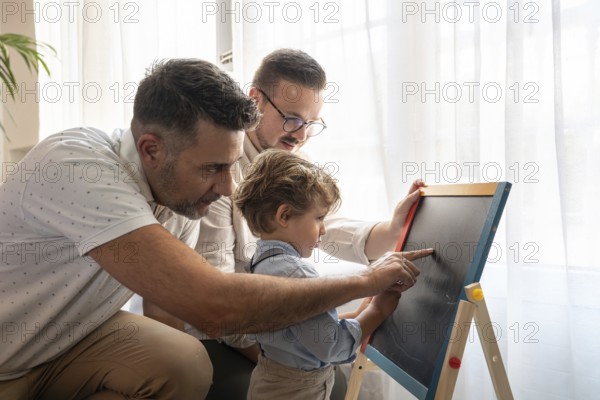 A loving gay couple engages with their young son, teaching him using a chalkboard. The warm,