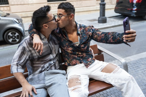 A latin gay couple shares a kiss while taking a selfie on a bench in Madrid. They display affection