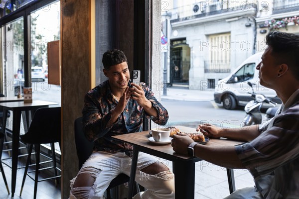 A latin gay couple enjoys coffee and pastries while sitting at a cafe. One partner is engaging with