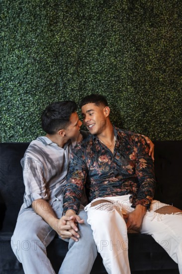 A loving latin gay couple sits closely on a sofa, holding hands and sharing a joyful, intimate
