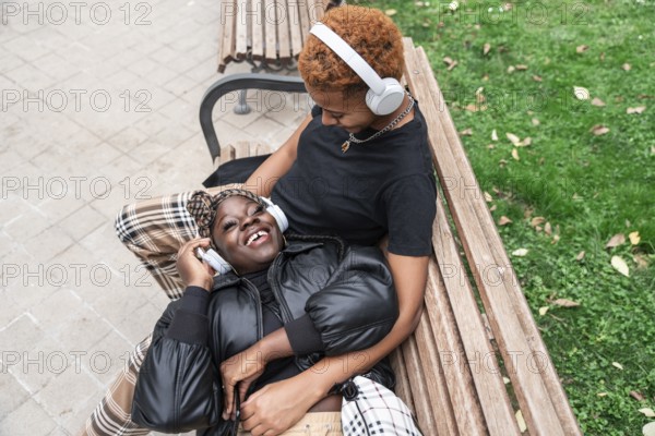 A joyful LGBT multiethnic couple relaxes on a park bench, listening to music through headphones.