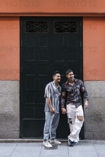 A joyful latin gay couple holds hands while standing in front of a colorful building in Madrid. The