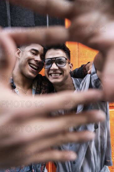 A joyful latin gay couple poses with smiles, framing their faces with their hands for a selfie