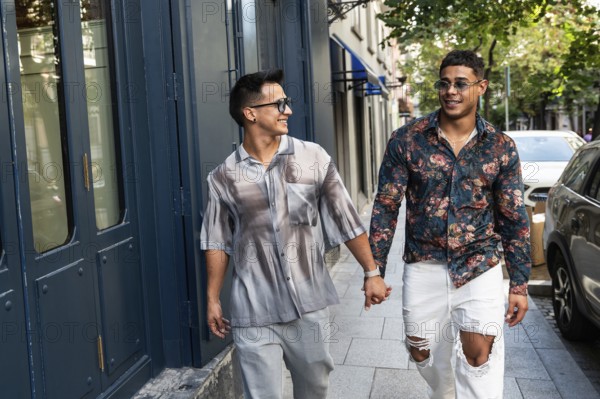 A happy latin gay couple holds hands while strolling down a lively city street in Madrid. They wear