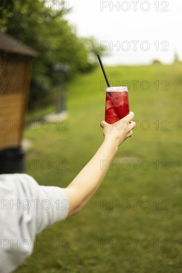A hand holding a refreshing red drink with ice and a straw, set against a lush green background on