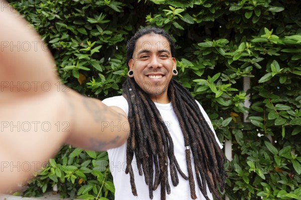 A cheerful man with long dreadlocks smiles while taking a selfie in a lush green garden in