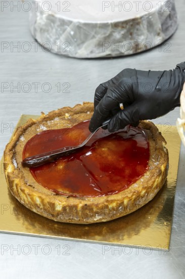 A baker wearing black gloves skillfully spreads glossy red glaze over a freshly baked tart The tart