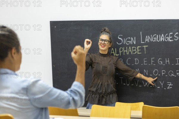 An instructor teaches sign language, pointing to an alphabet chart on a chalkboard A student