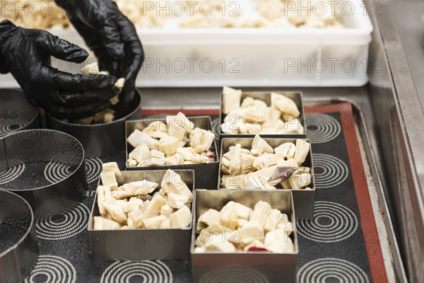 A baker wearing black gloves carefully arranges small pieces of dough in square molds on a baking