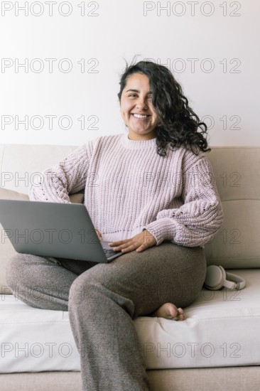 A cheerful young hispanic woman sits comfortably on a sofa at home, using her laptop She is dressed