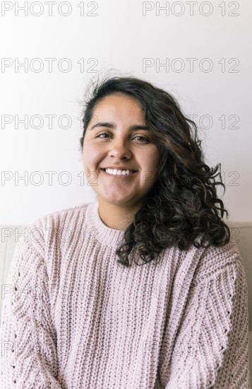 A young latin woman with curly hair smiles warmly, sitting indoors wearing a cozy sweater The