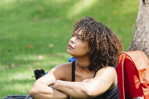 A Black woman sits under a tree in a Bogota park, resting her arms on her knees while gazing into