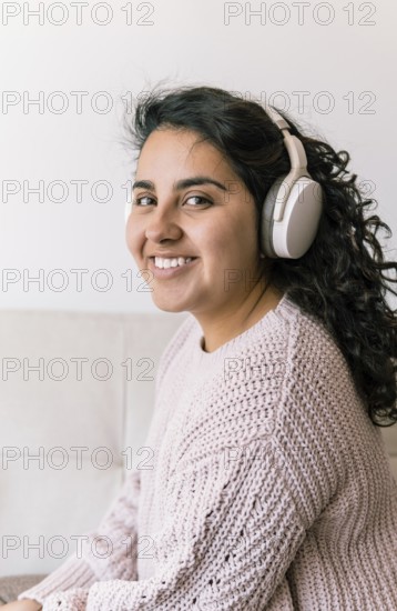 A young latin woman sits on a cozy couch, wearing headphones and a knitted sweater She is smiling,