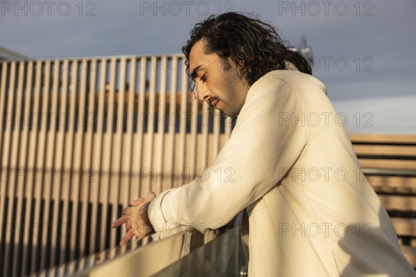 A man in a light shirt leans on a railing, captured in soft evening light His thoughtful expression