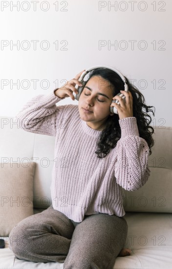 A young latin woman with curly hair relaxes on a couch at home, wearing a cozy sweater and